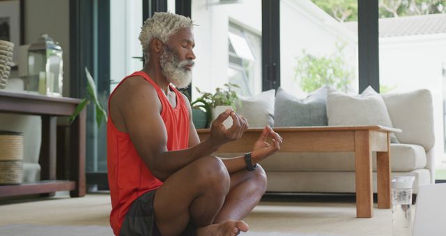 Elderly man sitting on floor of living room meditating. Appears focused, serene, and engaged in mindfulness practice. Ideal for topics about mental wellness, yoga routines, healthy aging, indoor activities, and promoting a balanced lifestyle.