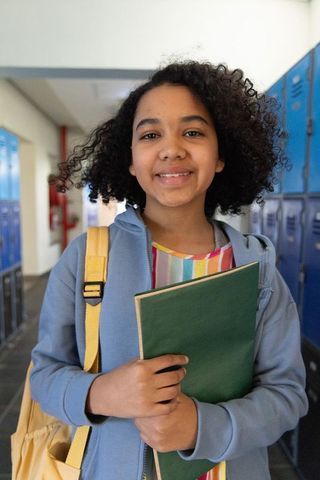 Smiling african american teen standing by lockers, holding green notebook, wearing yellow backpack