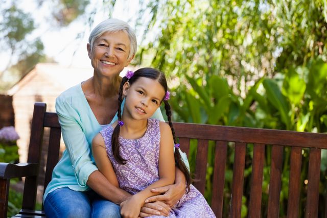 Portrait of smiling senior woman sitting with arm around girl on wooden bench at backyard
