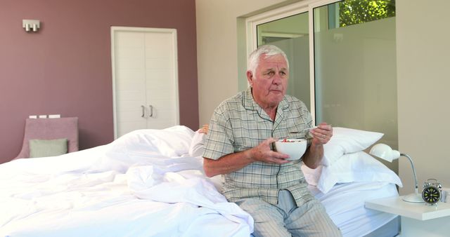 Elderly man eating breakfast in bed, wearing pajamas, in a modern and cozy bedroom. He looks calm and relaxed, enjoying his morning meal. Ideal for illustrating concepts of senior lifestyle, comfort, morning routine, home life, relaxation, and healthy habits.