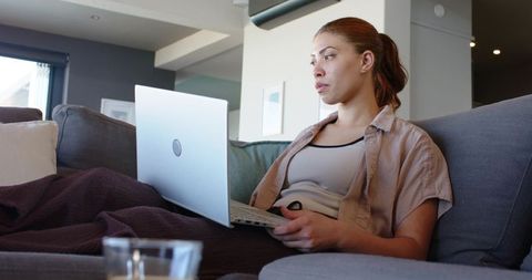 Young woman relaxing on couch using laptop, working from home with cozy blanket