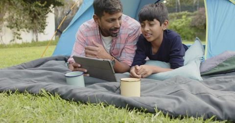 Father and Son Sharing Tablet While Camping on Gray Sleeping Pad by Blue Tent