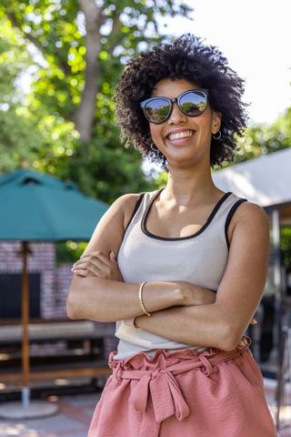 Confident african american woman smiling in sunglasses crossing arms on outdoor patio