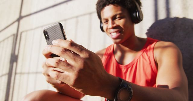 Young man in sleeveless red top and headphones, smiling while looking at smartphone outdoors. Great for illustrating themes of youth, technology, relaxation, summer activities or enjoying leisure time.