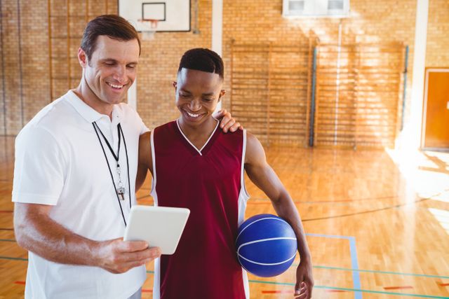Coach and basketball player standing in gym, reviewing strategy on tablet. Ideal for use in sports training, coaching, teamwork, and educational materials. Highlights use of technology in sports and mentor-athlete relationship.