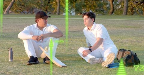Two cricketers sitting on grass talking during sunny break with bat and sport gear overlay