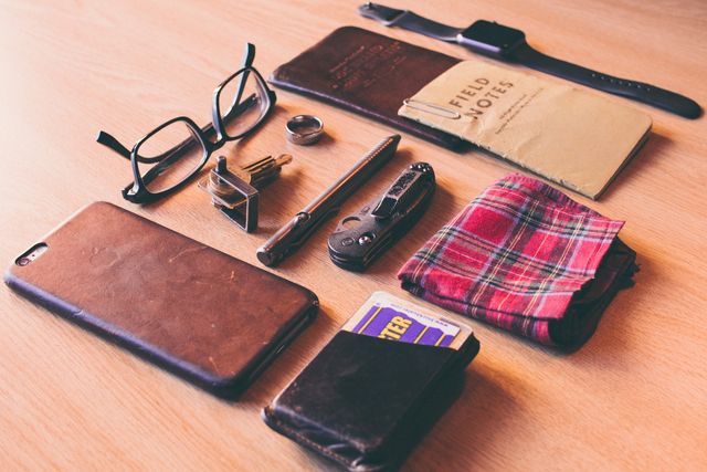 This image exhibits a variety of essential everyday carry items neatly arranged on a wooden desk. The items include glasses, a smartphone, wallets, a pocket notebook, a folded scarf, a multitool, a pen, a wristwatch, and a small stand. This image is ideal for websites and blogs discussing men's fashion, everyday carry essentials, personal organization, and lifestyle tips. It can also be used in advertising for EDC products, stationery, or men’s accessories.