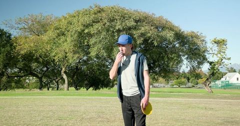 Man calling out holding yellow frisbee on park field wearing blue cap and vest