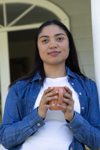 Indian woman on porch holding pink mug, wearing denim jacket and white top