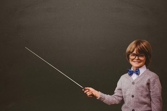 Child wearing glasses and bow tie holding pointer while smiling against chalkboard background. Ideal for use in educational materials, back-to-school promotions, or child learning environment concepts.