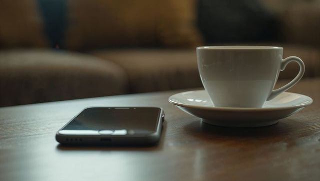 White ceramic coffee cup on saucer beside smartphone on wooden table in cozy lounge
