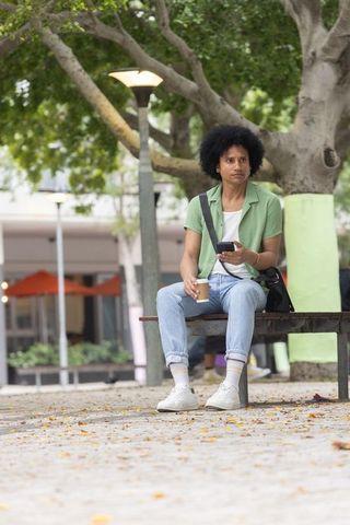 African american man sitting on bench holding takeaway coffee checking smartphone in urban plaza