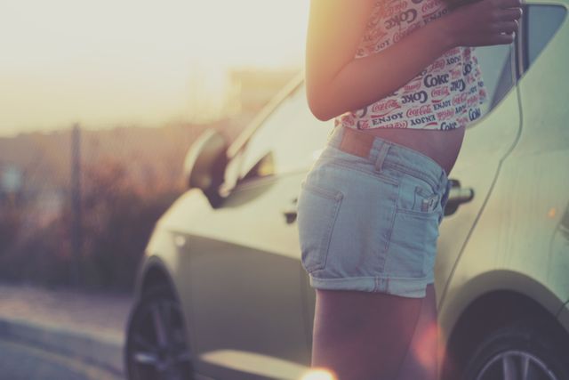 Close-up of a young woman standing by a car during sunset, wearing jean shorts and casual top. Ideal for advertisements, lifestyle blogs, and travel-related content.