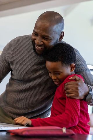 African american father embracing son while using laptop at table, bonding at home