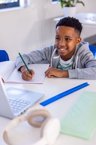 African american boy studying online smiling and writing at bright classroom desk