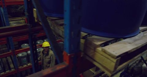 Warehouse worker inspecting pallet racking under stacked blue drums wearing yellow helmet