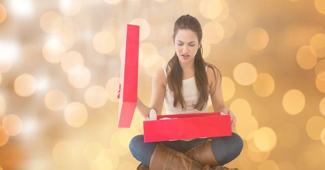 Young woman sitting casually, opening a red gift box with a disappointed expression against a warm bokeh background. This image can be used for themes related to holiday expectations, gift-giving, emotional reactions during holidays, or promotional materials for companies focusing on customer experience and satisfaction.