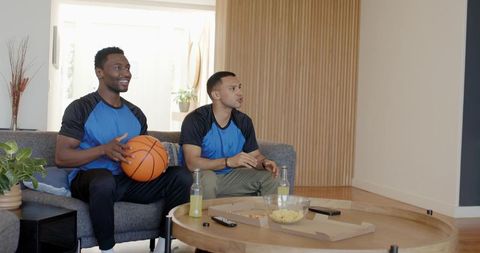Diverse male friends watching basketball on tv at home holding ball and snacking