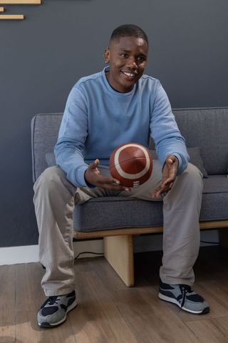 African american teen sitting on sofa holding basketball in modern minimalist living room