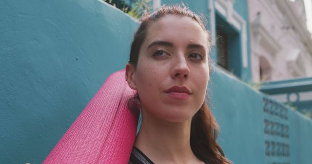 Young woman holding pink yoga mat on shoulder, standing against blue wall in outdoor urban environment. Ideal for promotions related to fitness, wellness, healthy lifestyles, and urban living. Useful for blogs, social media posts, advertisements, and websites focused on yoga, outdoor activities, or youth wellness.