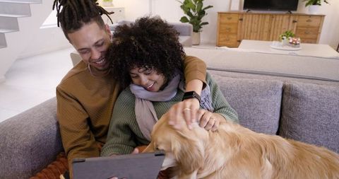 African american couple sharing tablet and cuddling golden retriever on cozy gray sofa