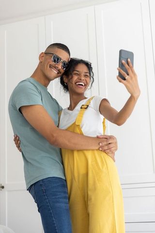 Young couple laughing and taking selfie at home, woman in yellow overalls