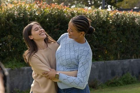 Two friends laughing and hugging in knit sweaters on sunny lawn, smartwatch visible