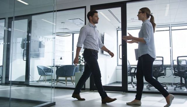 Walking coworkers chatting in modern glass-walled office corridor, business professionals