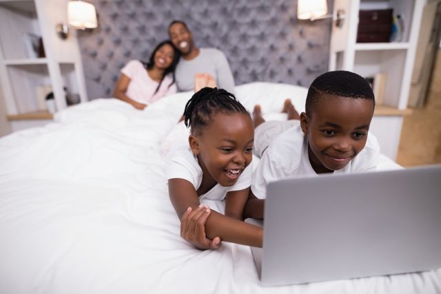 Happy siblings using laptop on bed with parents sitting in background at home