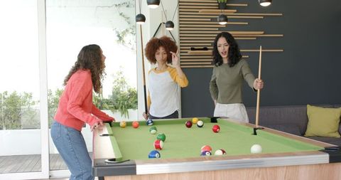 Diverse women playing pool, lining up cues and laughing in modern recreation room
