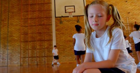 Young schoolgirl relaxing in gym with classmates playing basketball