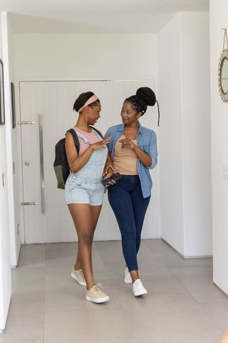 African mother and daughter walking through modern hallway holding smartphone and backpack