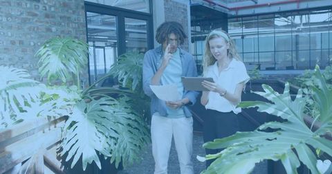 Young professionals collaborating using tablet and papers among lush plants in modern office atrium