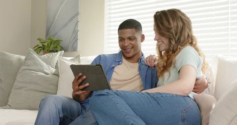 Diverse young couple relaxing on light sofa watching tablet and laughing together