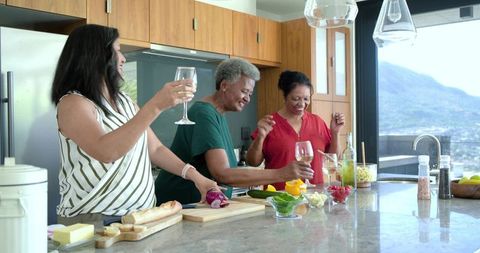 Friends cooking and laughing at modern kitchen island with wine, fresh produce, city view