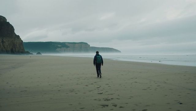 Solitary hiker walking along misty overcast beach with cliffs and trailing footprints