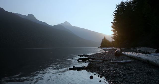 A serene lake at dusk, with the sun setting behind mountain peaks, creates a tranquil atmosphere. Driftwood and pebbles line the shore, accentuating the peacefulness of this natural setting.