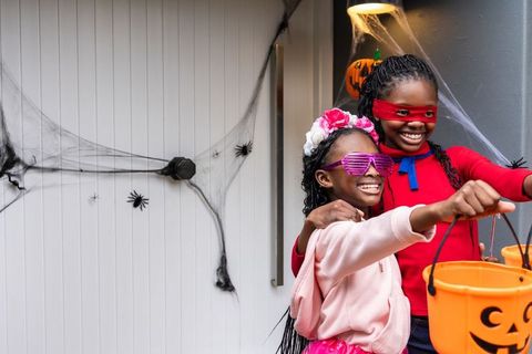 Joyful african american girls trick-or-treating in colorful costumes on spider-webbed porch