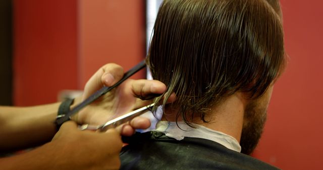 Barber carefully cutting man's hair with scissors in a barbershop with red walls. This image is useful for websites and blogs related to men's grooming and hairstyling, barbershop services, style and fashion magazines, and hair care tutorials and advertisements. Perfect for illustrating articles about professional haircut techniques and men's grooming trends.