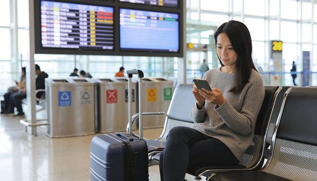 Young woman checking smartphone while waiting at airport gate with carry-on luggage
