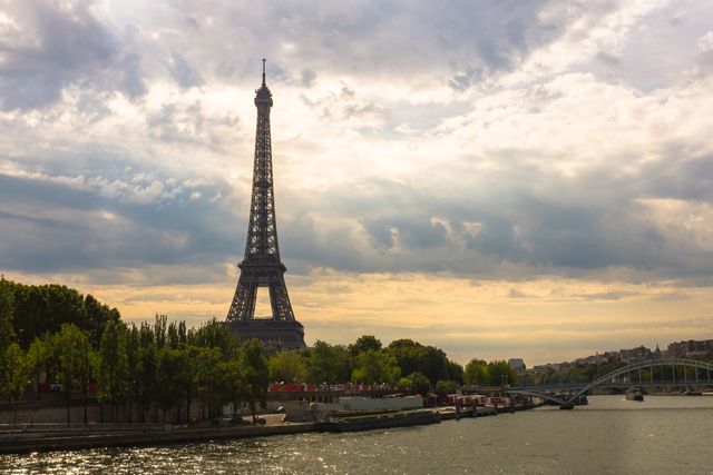 Eiffel Tower towering over the River Seine at sunset with partly cloudy sky and trees in the foreground, great for travel-related content, postcards, guidebooks, wallpapers, backgrounds, and web design.