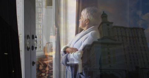 Senior woman in robe gazing through glass door with reflected city tower, contemplative morning