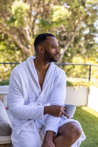 Relaxing mature african american man on sunlit patio in white robe holding mug