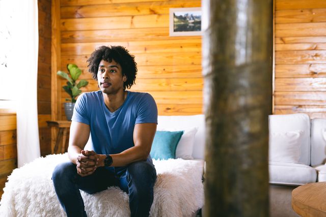 African American man sitting on a bed in a cozy log cabin with wooden walls. He is casually dressed in a blue t-shirt and jeans, looking thoughtful. This image can be used for lifestyle blogs, travel websites, or advertisements promoting relaxation, nature retreats, or cabin rentals.