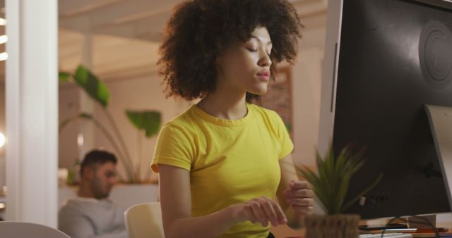 Young woman with curly hair and yellow shirt is working on a computer in a modern office. The workplace features a contemporary design with plants, contributing to a vibrant, productive atmosphere. Ideal for use in articles or materials related to modern offices, work environments, productivity, or technology.