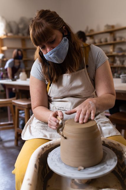 Caucasian female potter wearing face mask sitting by pottery wheel forming a dish. health and hygiene at pottery studio during coronavirus covid 19 pandemic.