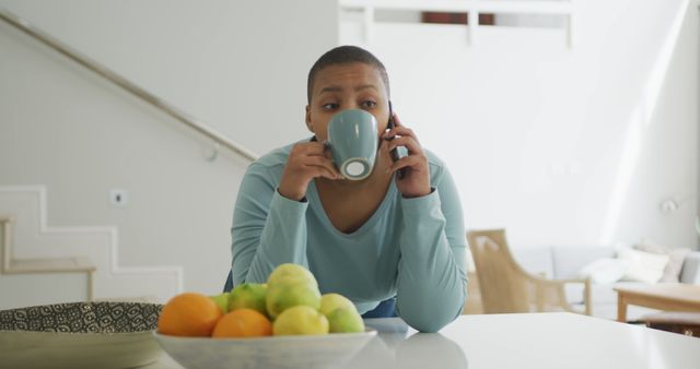 Woman leaning against kitchen counter, drinking from blue mug, multitasking with phone call. Background features bright, modern home with contemporary decor. Useful for themes related to daily routine, communication, and modern living.