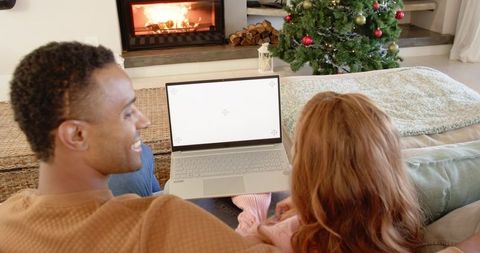 Diverse couple watching laptop by fireplace with christmas tree, cozy holiday evening