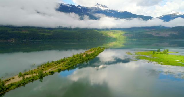 Aerial view displays a tranquil lake reflecting nearby mountains and clouds, with a thin strip of land featuring a hiking trail. Lush greenery edges the shoreline, enhancing the idyllic experience. Ideal for outdoor, travel, and adventure content. Perfect for backgrounds, travel blogs, and environmental inspirational imagery.