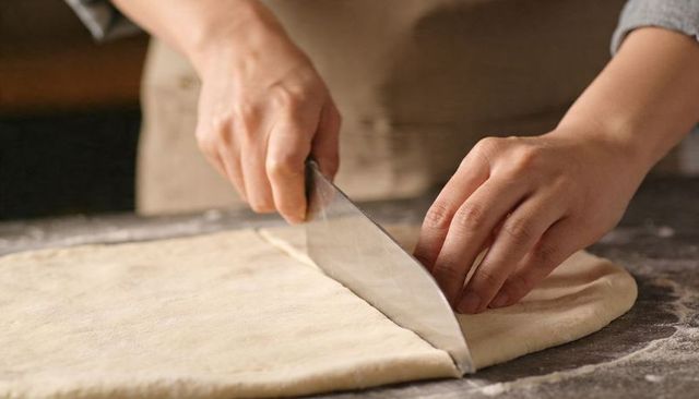 Cutting rolled dough with bench scraper on floured countertop preparing pastries for baking